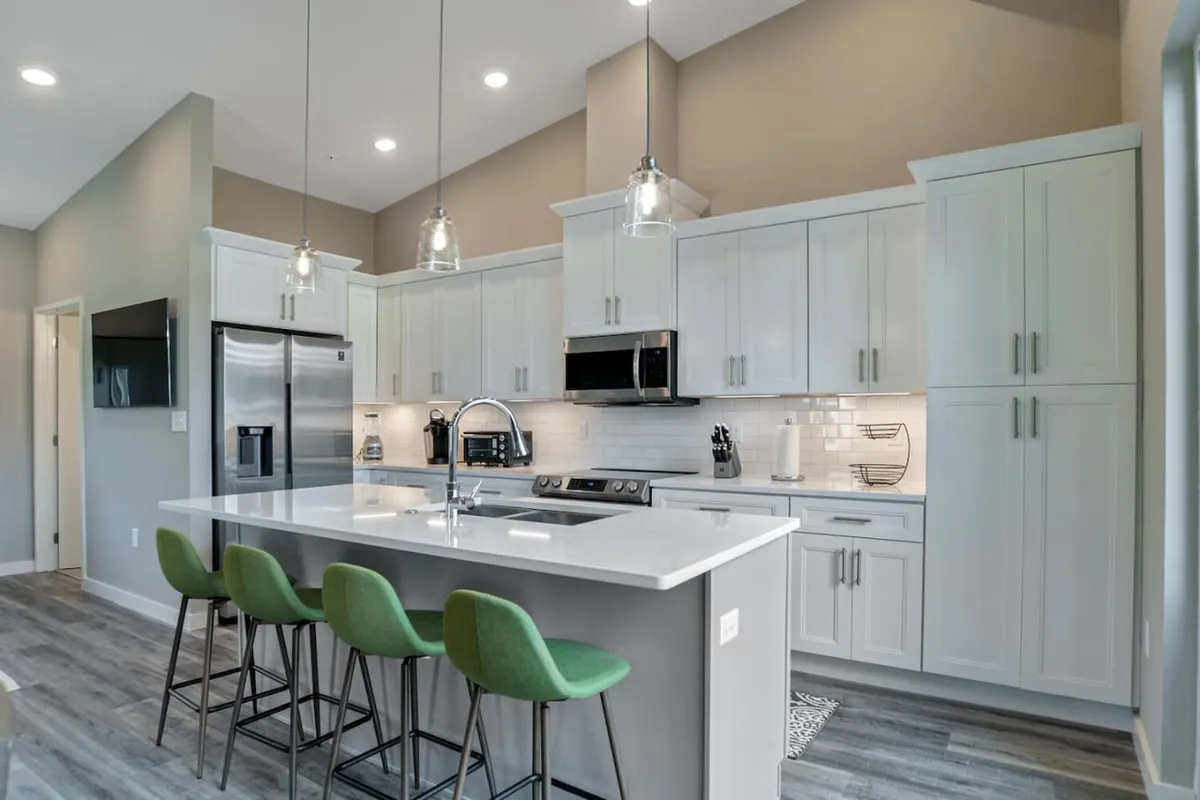 A spacious, modern kitchen featuring a large island with green bar stools, sleek white cabinetry, and stainless steel appliances. The room is filled with natural light, creating an inviting space for cooking or gathering. The island provides a casual dining spot and plenty of room for entertaining guests.