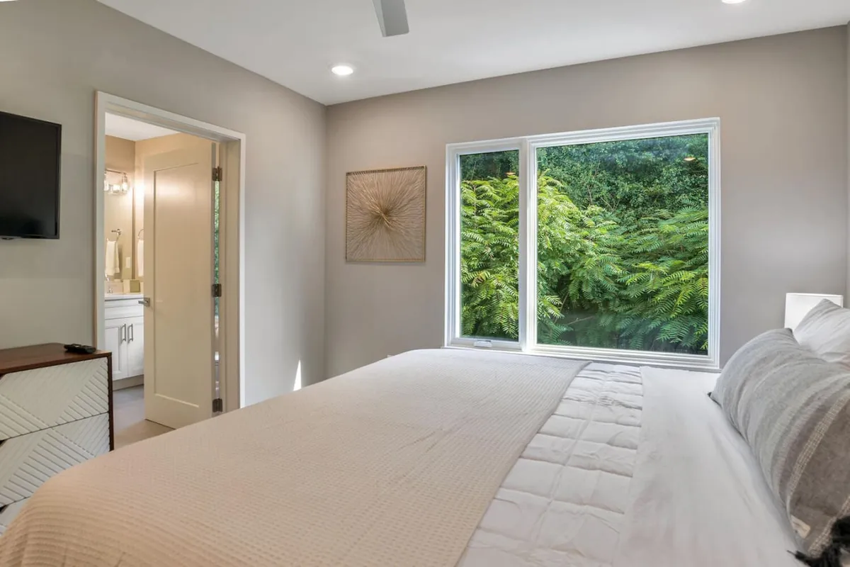 A cozy bedroom featuring a textured bed, a large window framing lush greenery outside, tasteful decorative art, and a wall-mounted TV. The space feels tranquil and connected to nature, ideal for unwinding after a day exploring downtown or the surrounding area.