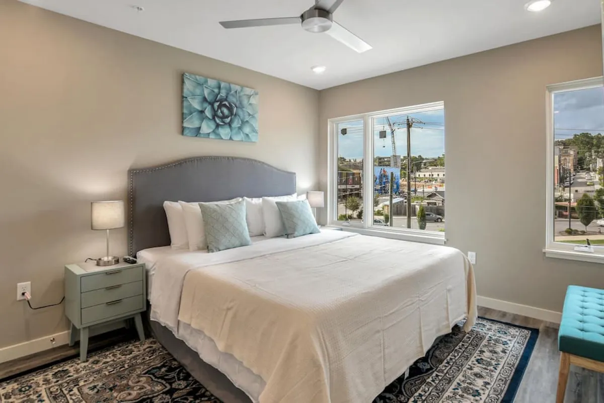 This image presents a serene bedroom featuring a plush bed, large windows that allow ample natural light, tasteful wall art, and cozy nightstands. The atmosphere is tranquil, suggesting a peaceful space for relaxation and privacy.