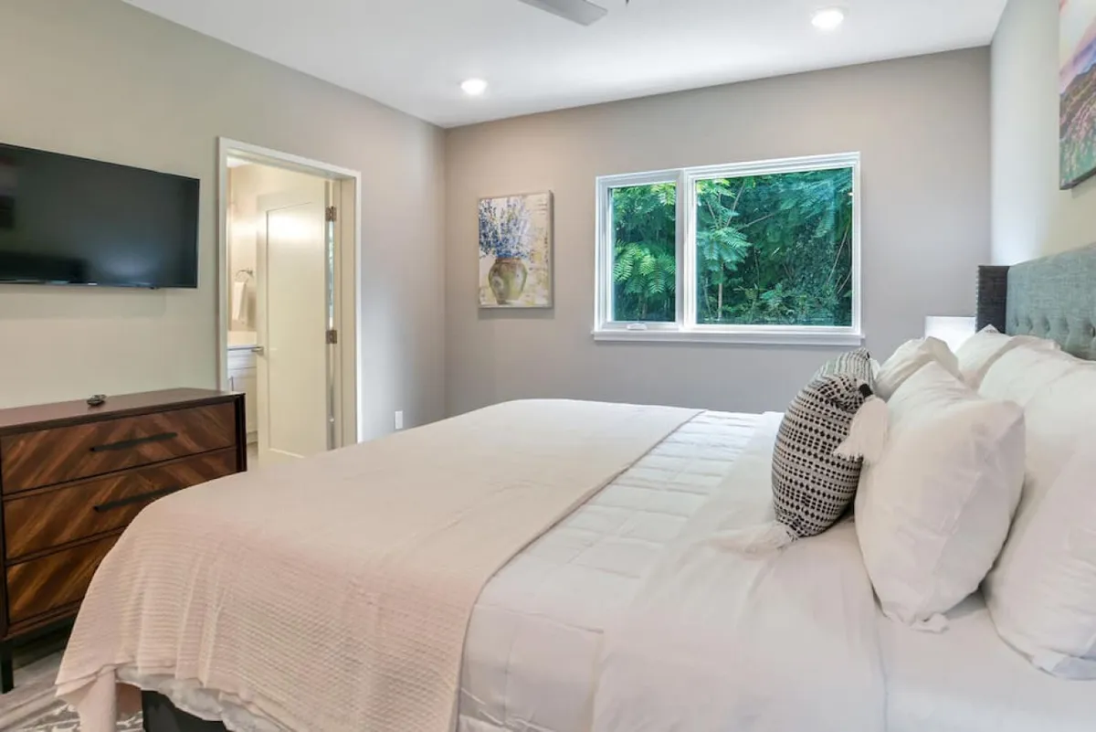 A bright, comfortable king bedroom with white linens, framed wall art, a large window with greenery outside, and a TV mounted on the wall. The space feels peaceful and offers both relaxation and entertainment options.