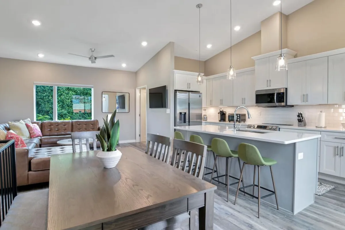 An open-plan living and dining area with a dining table, kitchen island, sofa, and expansive windows that let in abundant sunlight. The scene exudes warmth and togetherness, ideal for family meals, relaxed evenings, or socializing with friends.