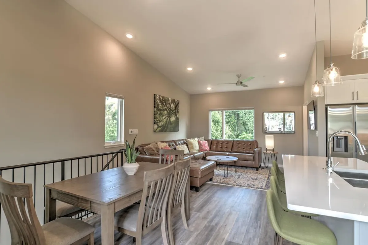 A spacious, light-filled open-plan living area featuring a dining table set for a group, a comfortable sofa, coffee table, and large windows that bring in natural light. Wall art and stylish decor create a welcoming atmosphere for social gatherings.
