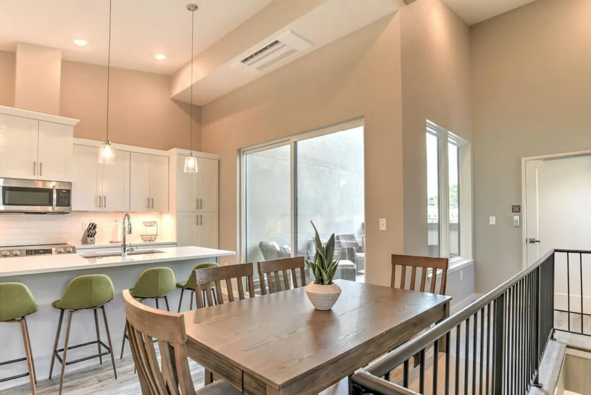 A modern kitchen and dining area with a central island, large windows, and a dining table set for multiple guests. The space is bathed in natural light, promoting a warm, communal vibe ideal for shared meals and entertaining.