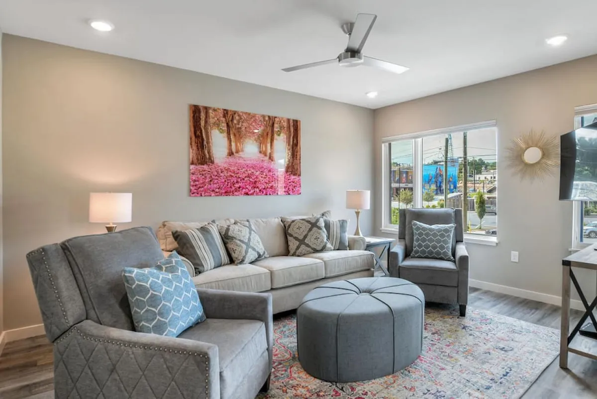 A warm and welcoming sitting area featuring a comfortable sofa, stylish accent chairs, a round ottoman, and large artwork depicting a tree-lined path. The space is bathed in natural light, inviting moments of relaxation, reading, or intimate conversation.
